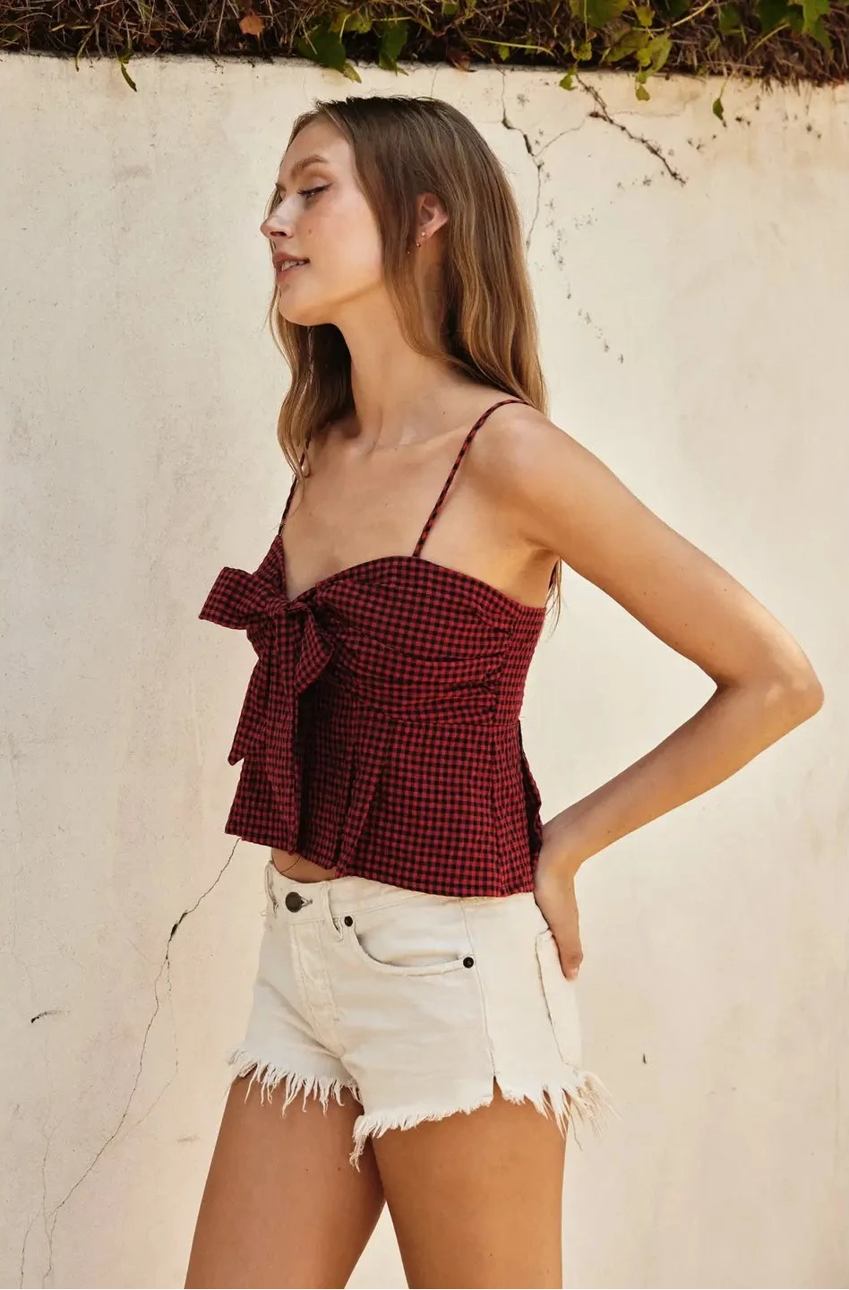 Woman wearing a red plaid top and white shorts against a beige wall.